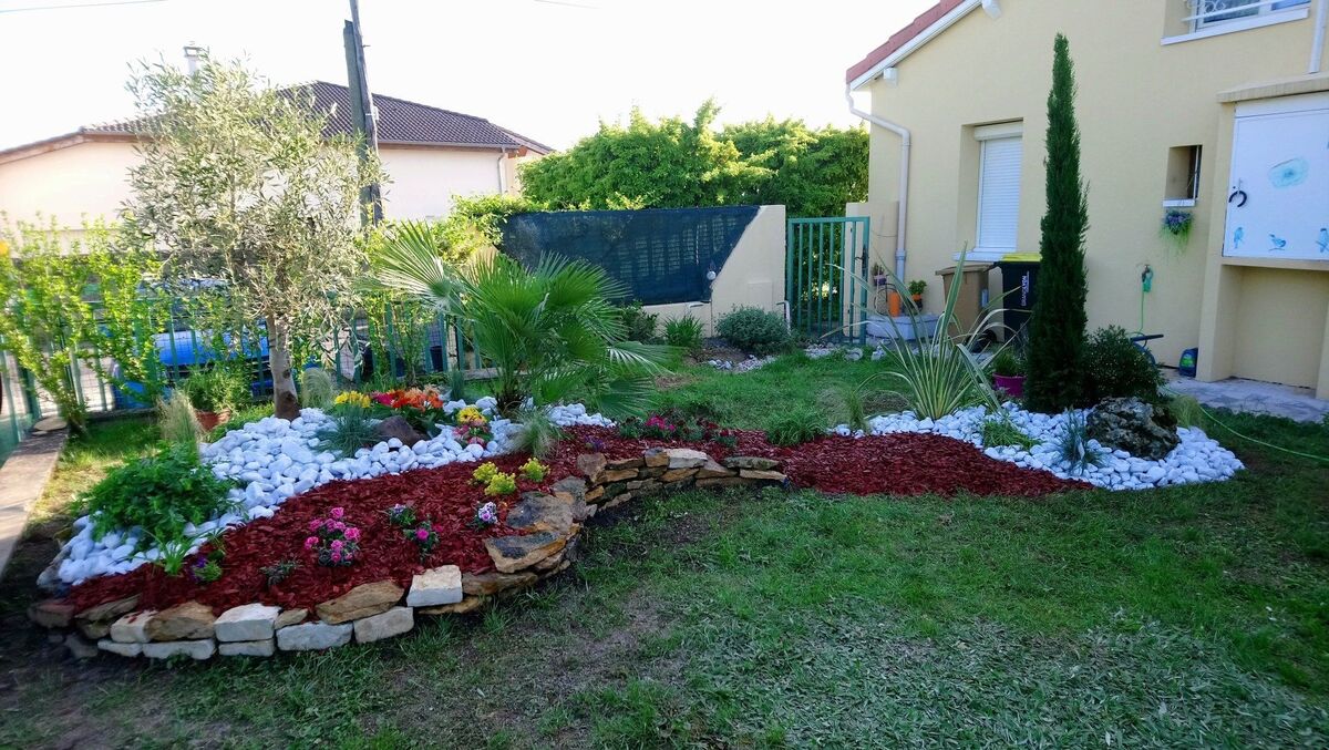 Pelouse bordée d'un massif de galets blancs, de paillage rouge et d'un cyprès
